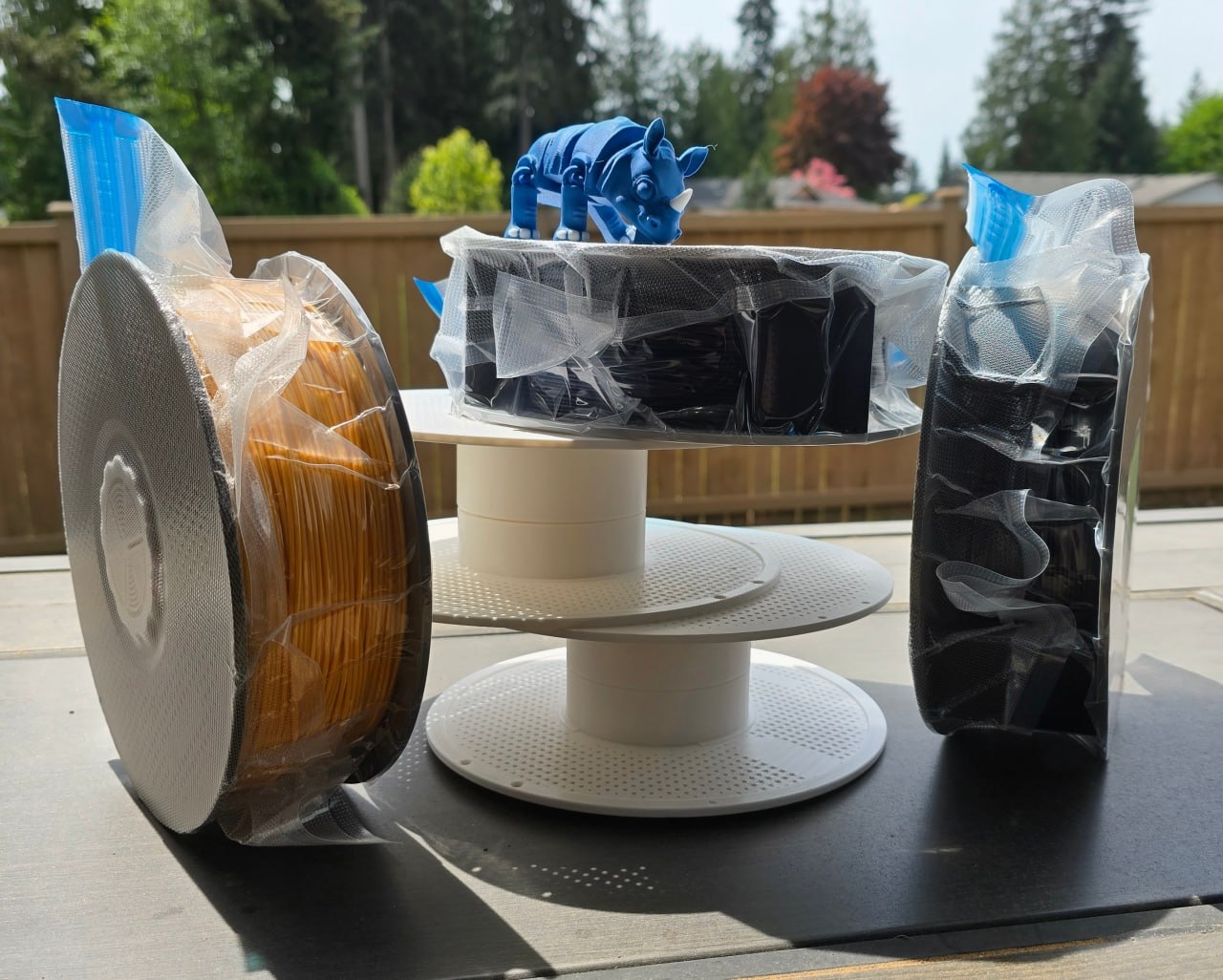 Three filament spools (PLA Silk gold, black ABS, black TPU) on a two-tier spool stand with a blue 3D printed rhino, outdoors